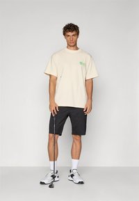 Bėriniško atspalvio trumpomis rankovėmis marškinėliai su žaliu grafiniu motyvu, derinami su juodais šortais. Modelis vilki baltas kojines ir sportinius batelius, laiko golfo lazdą.