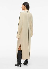 Robe tricotée beige à côtes avec des manches longues, des coutures latérales noires et de hautes fentes latérales. Portée avec des bottes noires à talons jusqu'aux genoux.