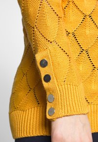 Stickad tröja i klar gul med ett diamantmönster, med ribbad mudd och tre metallknappar som accent på ärmen.