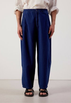 Personne portant un pantalon ample bleu foncé, une chemise blanche retroussée et des sandales noires à boucle, debout devant un fond uni.