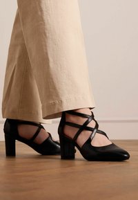 Scarpe con tacco alto in pelle nera, con punta affilata e design a strisce incrociate, posizionate su pantaloni beige chiaro su un pavimento di legno.