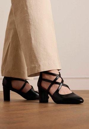 Scarpe con tacco alto in pelle nera, con punta affilata e design a strisce incrociate, posizionate su pantaloni beige chiaro su un pavimento di legno.
