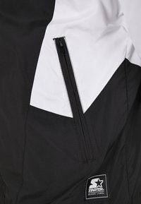 Section de veste noire et blanche avec une poche zippée verticale et un écusson logo Starter Black Label près du bas.