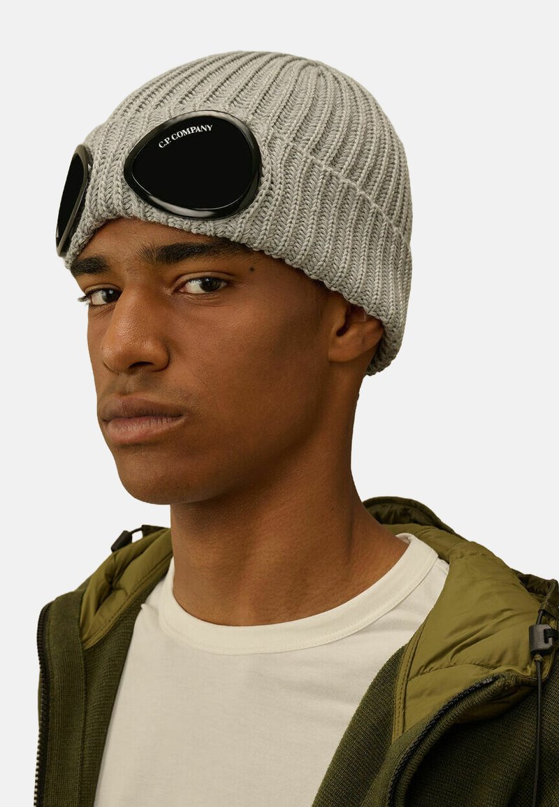 Beanie grigio a maglia con texture a coste, dotato di occhiali neri con un logo sul davanti. Indossato sopra una giacca verde e una camicia bianca.