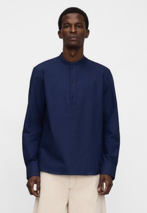 Uomo che indossa una maglia a maniche lunghe blu navy con una corta abbottonatura e pantaloni beige, in piedi davanti a uno sfondo bianco uniforme.