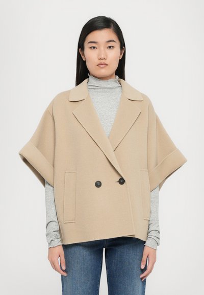 Beige dvouřadový plášť s kápí vyrobený z měkké látky, s velkými klopami, dvěma předními kapsami a černými knoflíky, nošený přes šedý rolák.