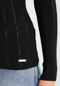 Top en tricot noir à côtes avec des découpes verticales, design ajusté, manches longues et une étiquette logo argentée en bas. Présente une surface texturée.
