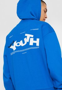 Modra kapuca z belim napisom na hrbtni strani, vključno z besedo "MLADOST". Narejena iz mehke tkanine z vrvico in sproščenim krojem.