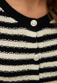 Zwart-witte gestreepte gebreide cardigan met gestructureerde zilveren knopen en een reliëfembleem bij de halslijn.