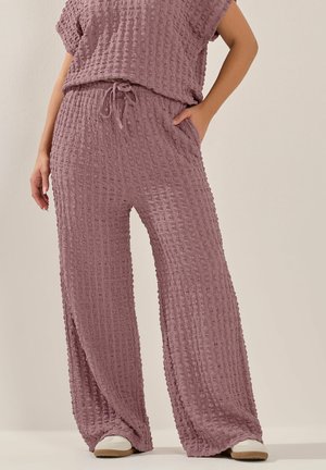 Kvinde iført tekstureret to-delt outfit i mauve med bukser med vide ben og hvide sneakers, stående med den ene hånd i lommen.