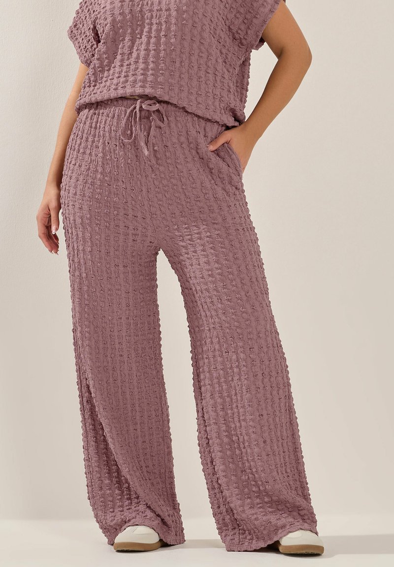 Frau trägt strukturiertes mauvefarbenes zweiteiliges Outfit mit weitem Bein und weißen Sneakers, steht mit einer Hand in der Tasche.