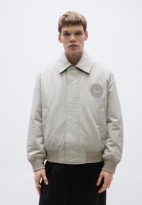 Ανοιχτό γκρι μπουφάν bomber με μεγάλο γιακά, ριμπ μανσέτες και λεπτομέρεια με μπαλώματα στο αριστερό στήθος. Με λεία υφή και άνετη εφαρμογή.