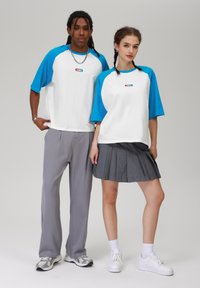 Valged T-särgid siniste raglanvarrukatega, hallid püksid ja hall plisseeritud seelik. T-särkidel on märkimisväärne logo detail. Mõlemal on jalas sneakers.
