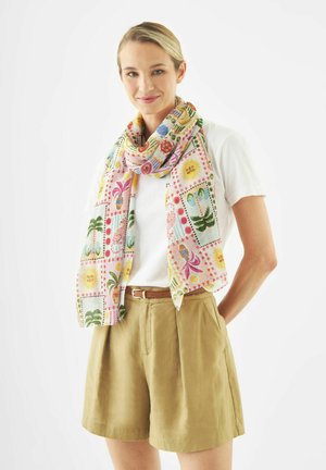 Jonge vrouw die een wit T-shirt draagt, beige short met riem en een kleurrijke sjaal met zon-, palmboom- en fruitpatronen, glimlachend met haar handen achter haar rug.