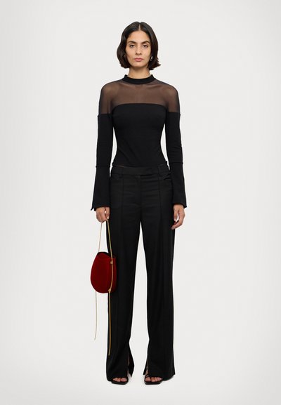 Haut long à manches transparentes noir avec un col haut, associé à un pantalon noir ajusté avec des fentes sur les côtés et des sandales noires. Sac à main rouge en accent.