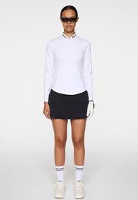 Haut blanc à manches longues avec col et accents noirs, associé à une mini-jupe côtelée noire. Des baskets blanches et des lunettes de soleil sportives complètent le look.