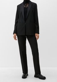 Abito nero composto da un blazer sartoriale con revers in satin e pantaloni dritti. L'outfit include un top in pizzo nero sotto.