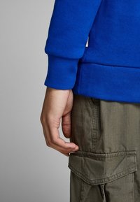 Mano maschile rilassata al lato, indossando una camicia a maniche lunghe blu e pantaloni cargo verde oliva con tasca a pattina.