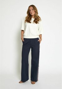 White cropped sweatshirt and navy pants with side stripe details; model poses with hands in pockets, wearing brown sandals.