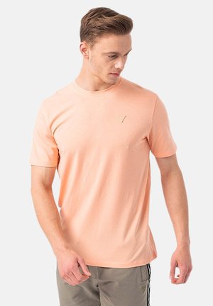 Homme portant un t-shirt pêche à manches courtes et un pantalon gris, regardant vers le bas et légèrement sur le côté sur un fond blanc uni.