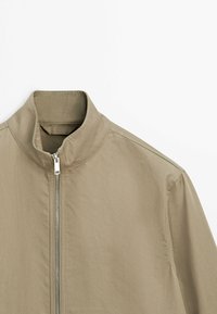 Giacca marrone chiaro con collo alla coreana e cerniera argento, vista frontalmente su sfondo bianco.