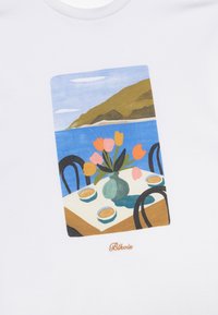 T-shirt blanc avec une illustration colorée d'une table avec un vase de tulipes et des tasses, sur fond de paysage côtier.
