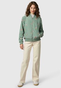 Kobieta o kręconych włosach, ubraną w zieloną bomberkę w kwiaty, beżowe spodnie o szerokich nogawkach oraz białe trampki, stojąca na neutralnym tle.