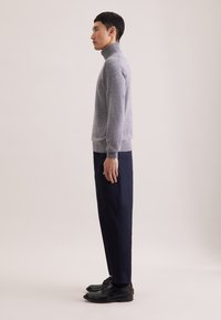 Grauer Rollkragenpullover aus Strickmaterial, kombiniert mit navyblauen Hosen und schwarzen Schuhen. Das Modell posiert im Profil vor einem hellen Hintergrund.