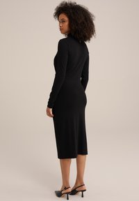 Schwarz langärmliges Kleid mit figurbetonter Silhouette, knielangem Saum, weichem Material und einem Rückenausschnitt. Getragen mit schwarzen Absatzschuhen.