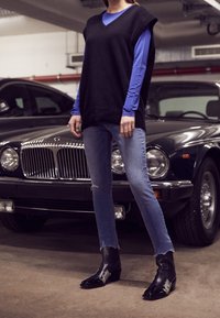 Suéter negro de punto sin mangas sobre una camisa de manga larga azul, combinado con jeans desgastados de color azul claro y botas negras hasta el tobillo con toques metálicos.