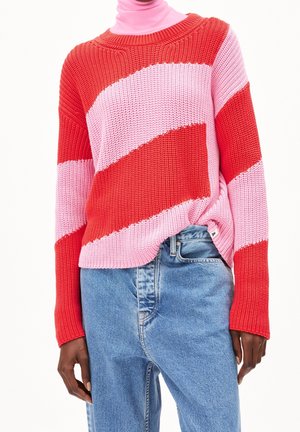 Personne portant un pull en maille rayé diagonal rouge et rose sur un col roulé rose, associé à un jean en denim bleu taille haute.