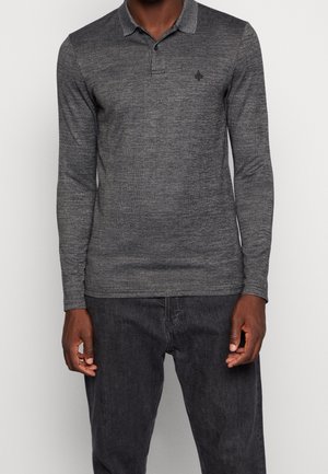 Mężczyzna ubrany w dopasowaną, długą, ciemnoszarą koszulkę polo w kratę oraz czarne jeansy, stojący na tle jednolitego jasnego tła.