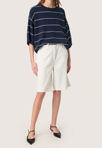 Oversized navyfarbener Strickpullover mit weißen horizontalen Streifen, kombiniert mit weißen knielangen Shorts und schwarzen spitz zulaufenden Ballerinas.