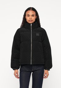 Czarna ocieplana kurtka z teksturowanym geometrycznym wzorem, wysoki kołnierz, zamek błyskawiczny i ściągacze przy mankietach. Nosi się ją na ciemnych dżinsach.