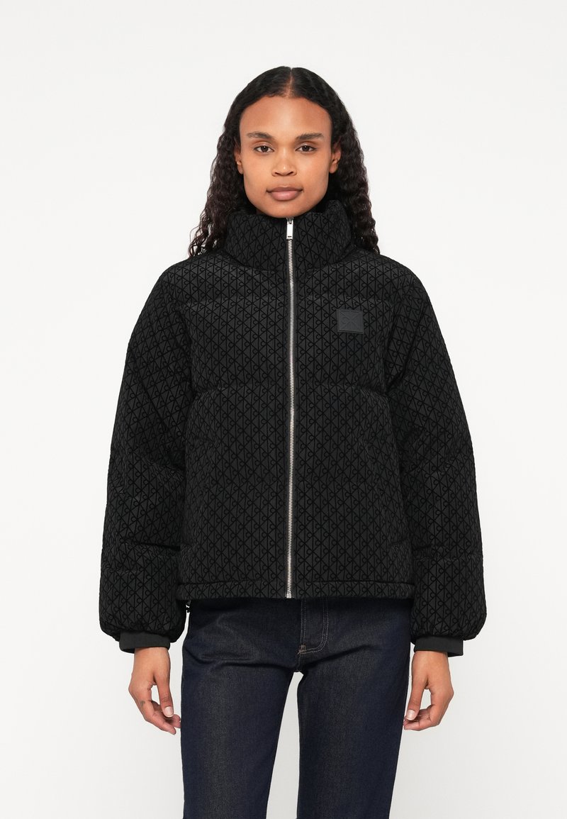 Czarna ocieplana kurtka z teksturowanym geometrycznym wzorem, wysoki kołnierz, zamek błyskawiczny i ściągacze przy mankietach. Nosi się ją na ciemnych dżinsach.