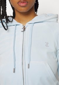 En person med flätat hår bär en ljusblå zip-up hoodie med en silverfärgad dragkedja och en liten prydnadslogotyp på bröstet.