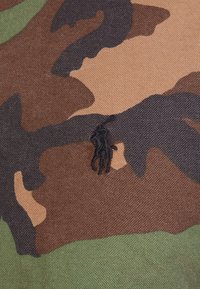 Tela de camuflaje que muestra tonos de marrón, verde y negro, con un pequeño logo bordado de un jugador de polo en negro centrado en el diseño.