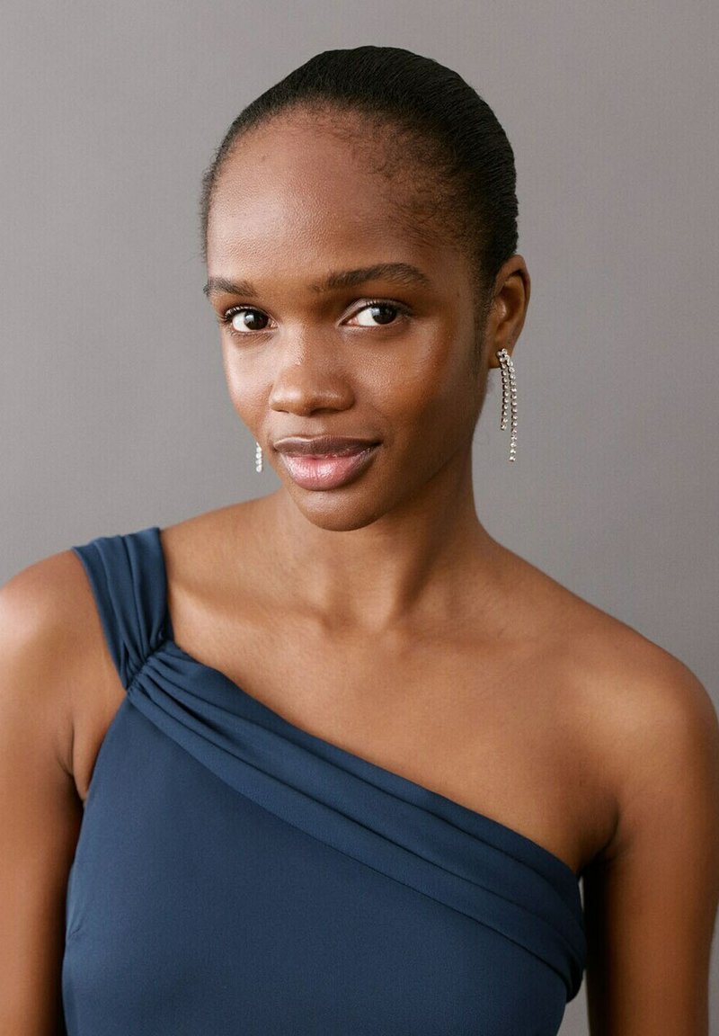 Mlada ženska s temno poltjo in lasmi zavezanim nazaj, oblečena v temno modro enoramno majico ter z dolgimi visečimi uhani, na sivem ozadju.