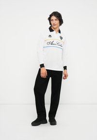 Camisa polo de manga larga blanca con collar negro, con rayas azules y amarillas y bordado. Combinada con pantalones negros y zapatos negros.