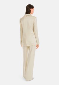 Beige tailored suit with a single-back vent and subtle sheen. Long sleeves, wide-leg trousers, single button detail at the back of the blazer.