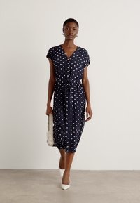 Navy dress with white polka dots, short sleeves, V-neck, cinched waist, and midi length. Worn with white shoes and holding a light handbag.