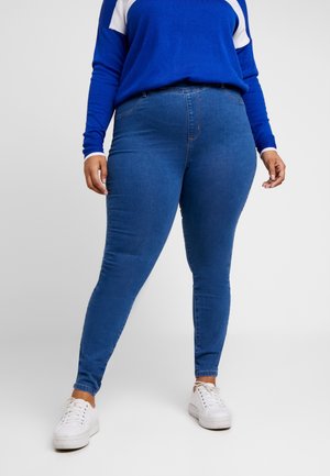 Personne portant un jean skinny bleu, des baskets blanches et un haut à manches longues bleu avec des accents blancs sur les épaules, debout devant un arrière-plan uni.