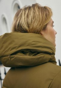 Žena s blond vlasy má na sobě olivově zelenou zateplenou bundu s velkou kapsou na zip a stojí nakloněná k jedné straně na pozadí rozmazané přírody.