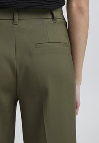 Olivově zelené kalhoty s hladkým povrchem, s zadní kapsou a poutky na opasek, viděné zezadu. Čisté linie a dobře padnoucí design.