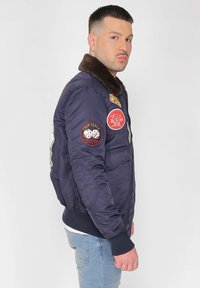 Marineblauer Bomberjacke mit Fellkragen, versehen mit verschiedenen Patches einschließlich roter und gelber Akzente, Reißverschluss und gerippten Bündchen und Saum.