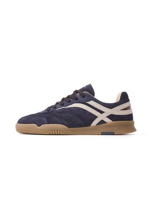 Donkerblauwe suède sneaker met beige strepen, bruine rubberen zool en zwarte veters, zijaanzicht op een witte achtergrond.