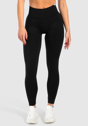 Leggings neri a vita alta realizzati in tessuto elasticizzato, con una texture liscia e un design aderente, abbinati a scarpe da ginnastica bianche.