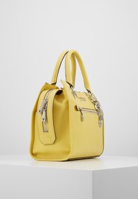 Keltainen tekstuurilla varustettu nahkabag, jossa on kaksinkertaiset kahvat, etupuolella vetoketjullinen tasku ja hopeanvärisiä vetoketjusopia valkoisella pinnalla.