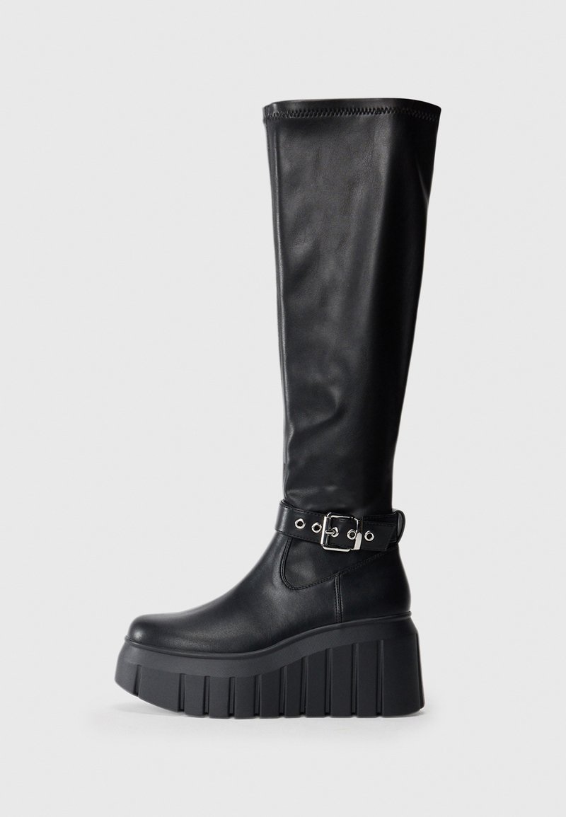 Botas pretas até ao joelho feitas de couro liso, com uma sola plataforma grossa, um salto robusto e um detalhe de cinto ajustável perto do tornozelo.