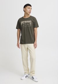 Oliven grønn bomulls t-skjorte med et grafisk trykk som sier "Original Global Jeansmaker," sammen med lyse beige bukser og hvite joggesko.